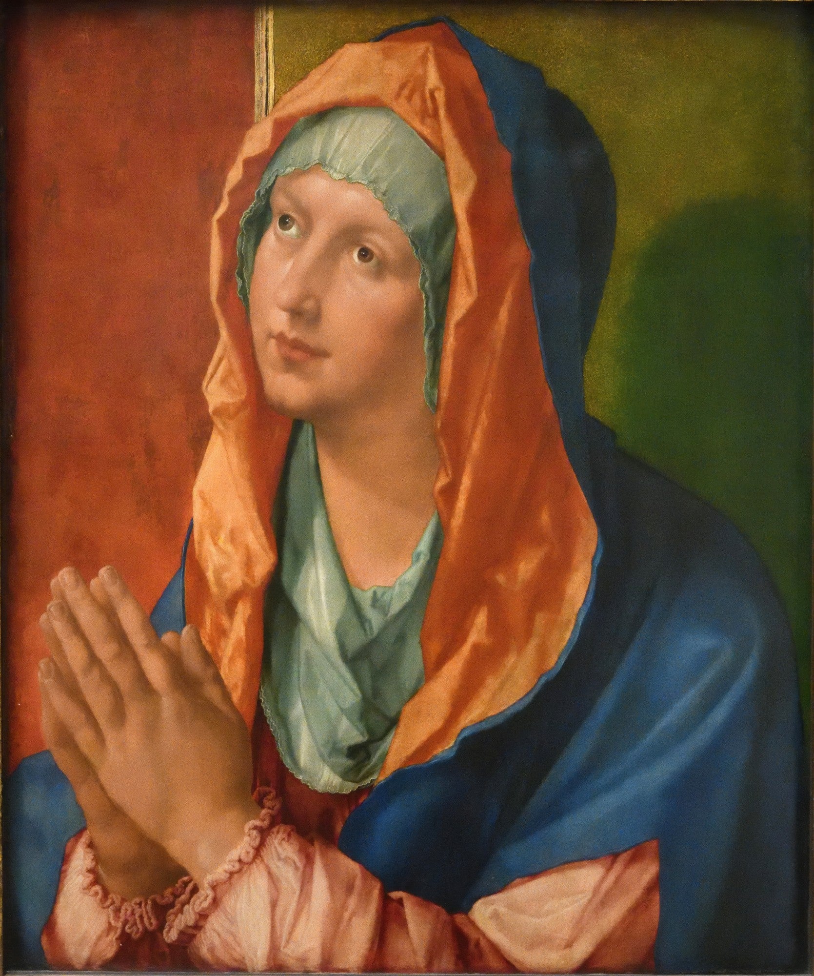 Quadro decorativo da Virgem Maria em oração, atribuída a Albrecht Dürer, com manto azul, véu laranja e fundo verde e vermelho. As mãos estão postas em prece e o olhar está elevado.