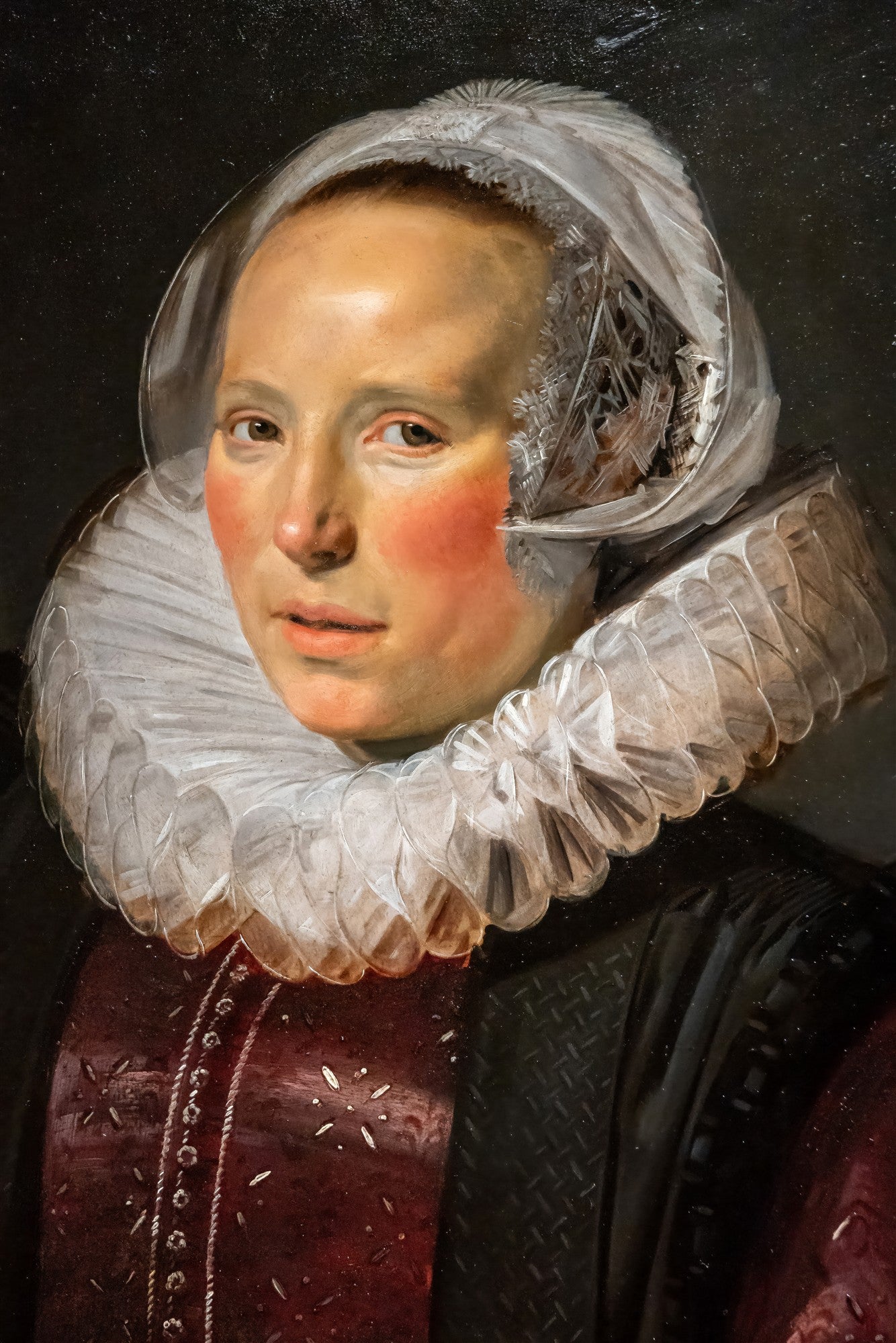 Retrato de uma mulher do século XVII, atribuído a Frans Hals, com touca branca e gola de renda frisada. A mulher tem pele clara, bochechas rosadas e veste um corpete escuro com detalhes em vermelho bordô. O fundo é escuro e liso, destacando a figura central.