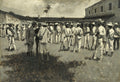 Pintura a óleo de Frederic Remington, 'Cuban Emigrès', mostrando um grupo de homens, alguns em uniformes militares, outros em trajes civis, reunidos em um pátio, com edifícios ao fundo e palmeiras. Predominam tons de cinza, branco e preto.