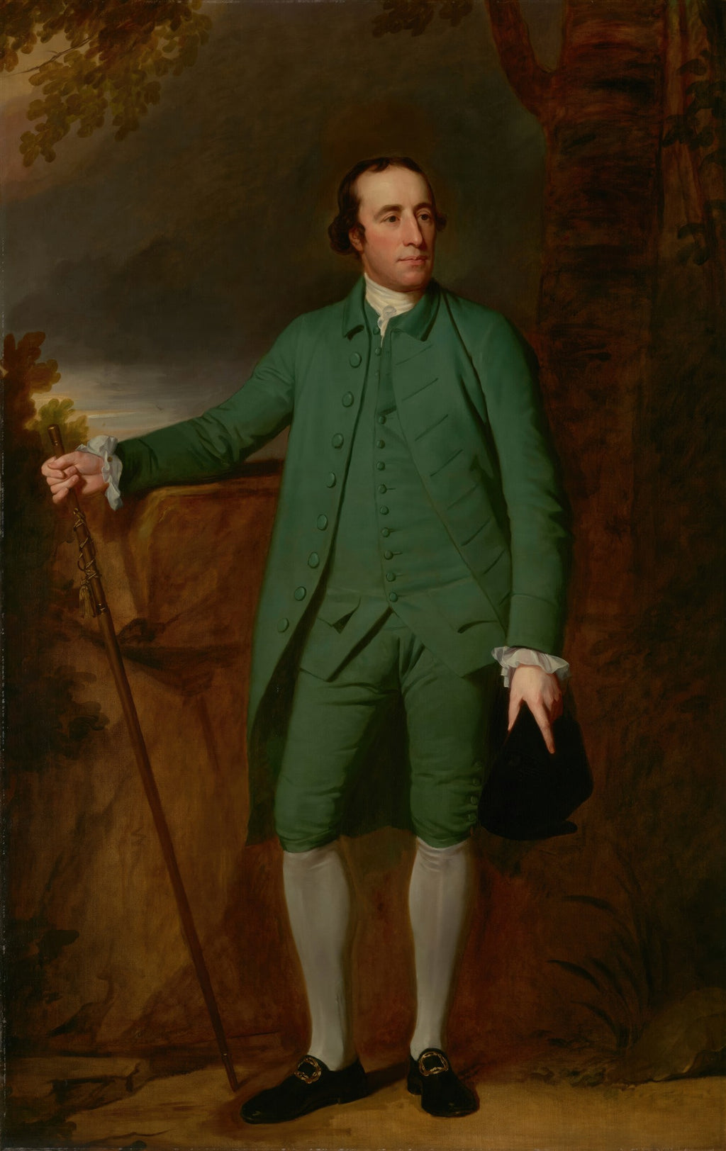 Retrato de um cavalheiro elegante em pé, vestido com um traje verde escuro, segurando um chapéu preto na mão direita e apoiado em uma bengala com a mão esquerda, em um cenário de paisagem em tons terrosos. Atribuído a George Romney.