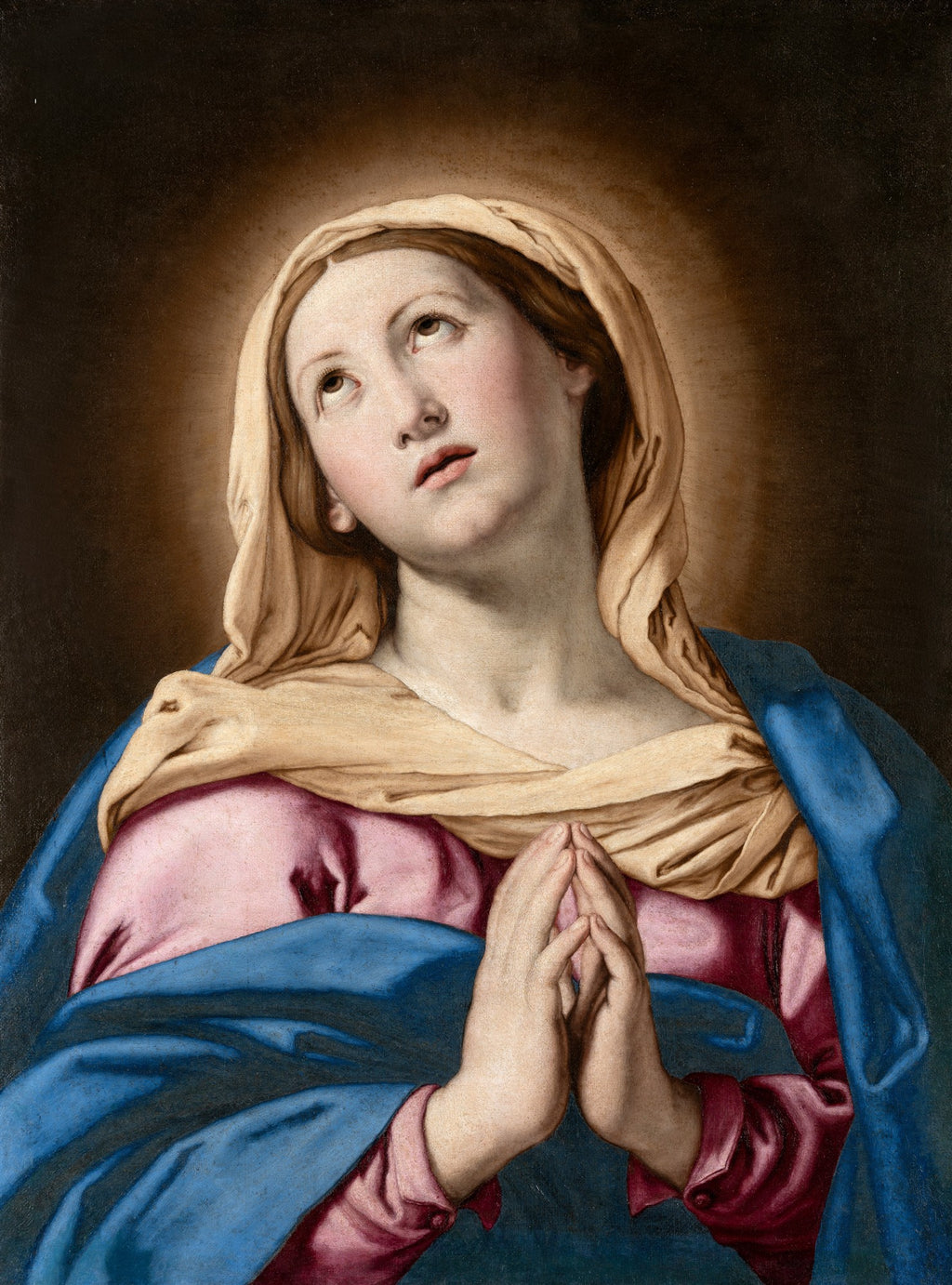 Pintura a óleo de 'A Virgem em Oração' de Giovanni Battista Sassoferrato. Retrata a Virgem Maria com as mãos postas em oração, olhando para cima com expressão serena. Ela veste um manto azul sobre uma túnica rosa e um véu bege. O fundo é escuro com um halo de luz ao redor da cabeça.
