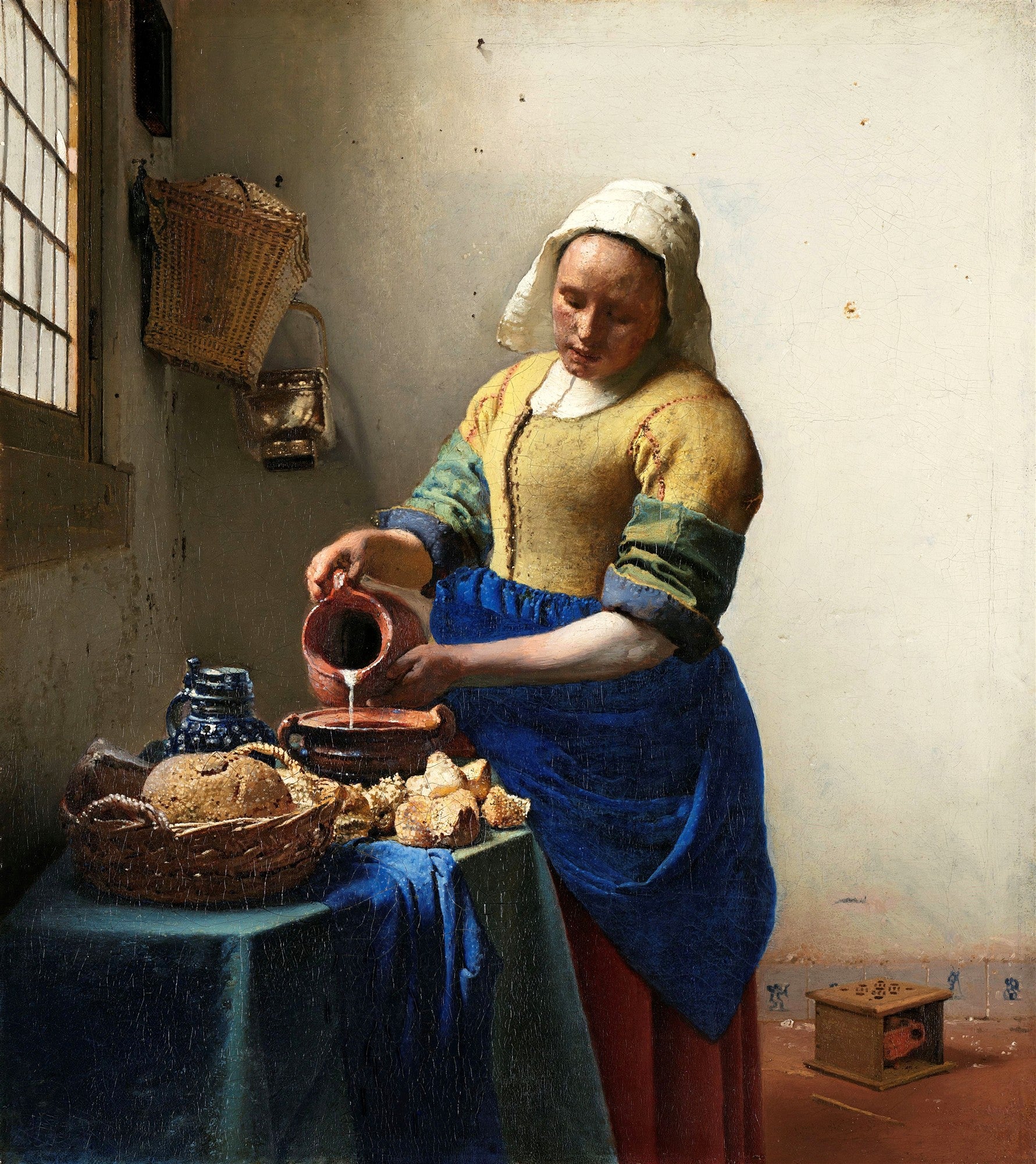 Uma mulher vestida com roupas de época, derramando leite de uma jarra para uma tigela sobre uma mesa com pães e cestas, em uma cena de interior iluminada por uma janela. Obra 'A Leiteira' de Johannes Vermeer.