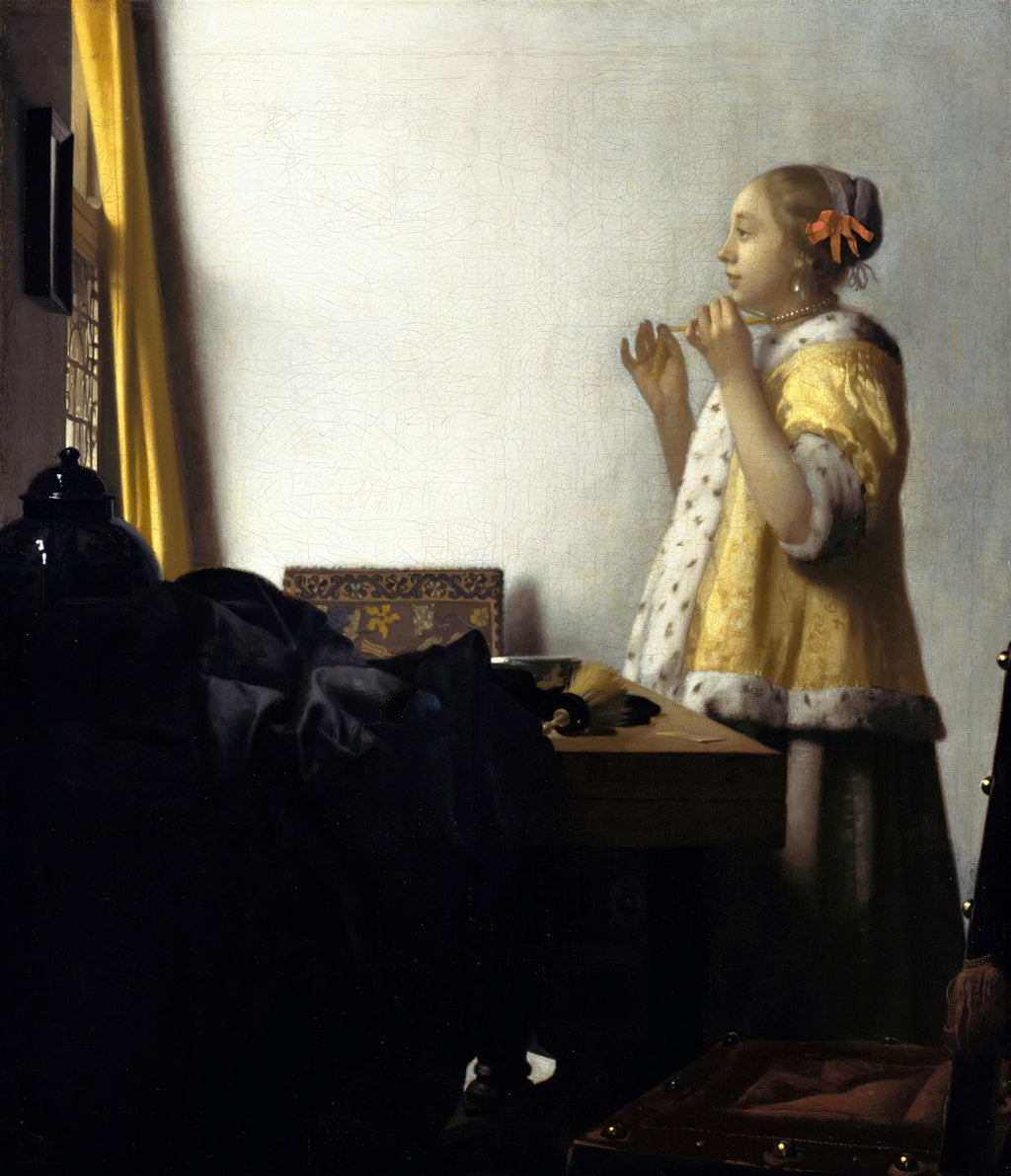 Quadro decorativo 'A Mulher com o Colar de Pérolas' de Johannes Vermeer, mostrando uma jovem mulher em um quarto iluminado pela janela, ajustando um colar de pérolas, vestindo um robe amarelo com acabamento de pele branca. A luz incide sobre a parede clara e o rosto da mulher, enquanto o lado esquerdo do ambiente está em sombra, com uma cortina amarela e um objeto escuro sobre uma mesa. A imagem transmite uma sensação de tranquilidade e intimidade.