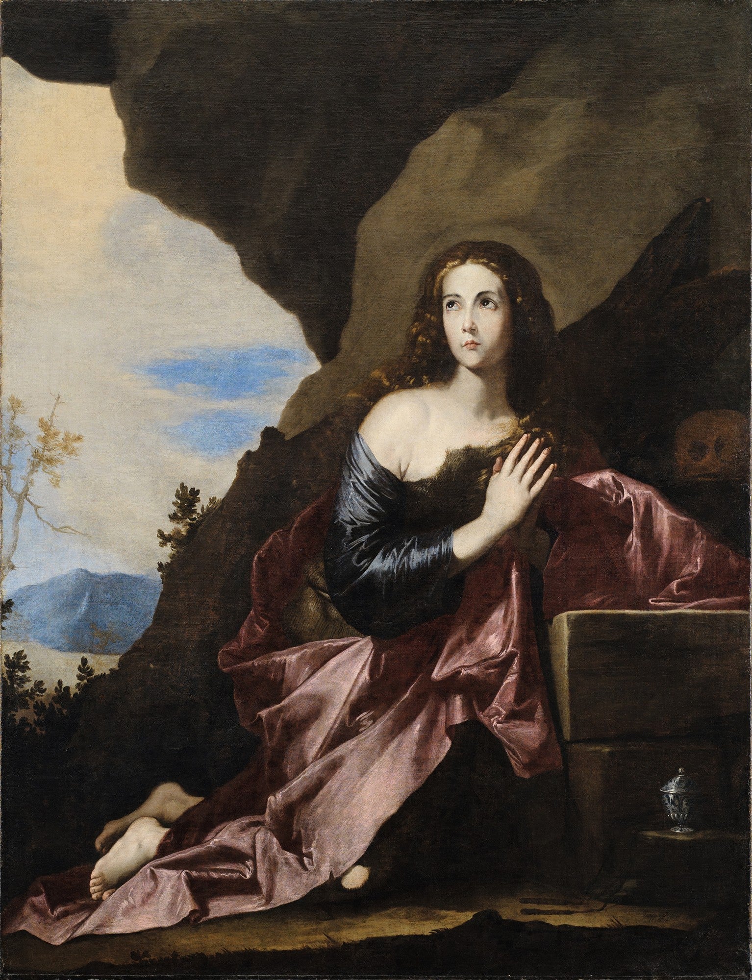 Quadro decorativo mostrando Maria Madalena penitente, ajoelhada em uma caverna escura, com as mãos em oração e o olhar para cima. Ela veste um manto azul escuro e um tecido rosado, com um crânio à sua direita. A cena é dramática e iluminada por uma luz vinda do canto superior esquerdo.