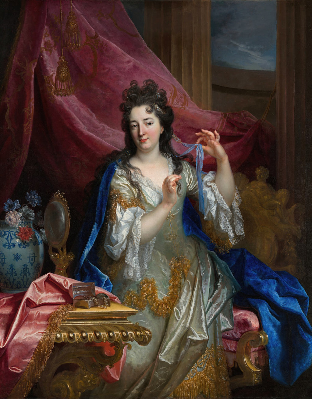 Retrato de uma mulher elegante sentada em um ambiente luxuoso, vestindo um vestido de seda claro com detalhes dourados e um manto azul, segurando uma fita azul. A mulher possui cabelos escuros e cacheados e seu olhar é direto. Ao lado, uma mesa com espelho e caixa de joias.