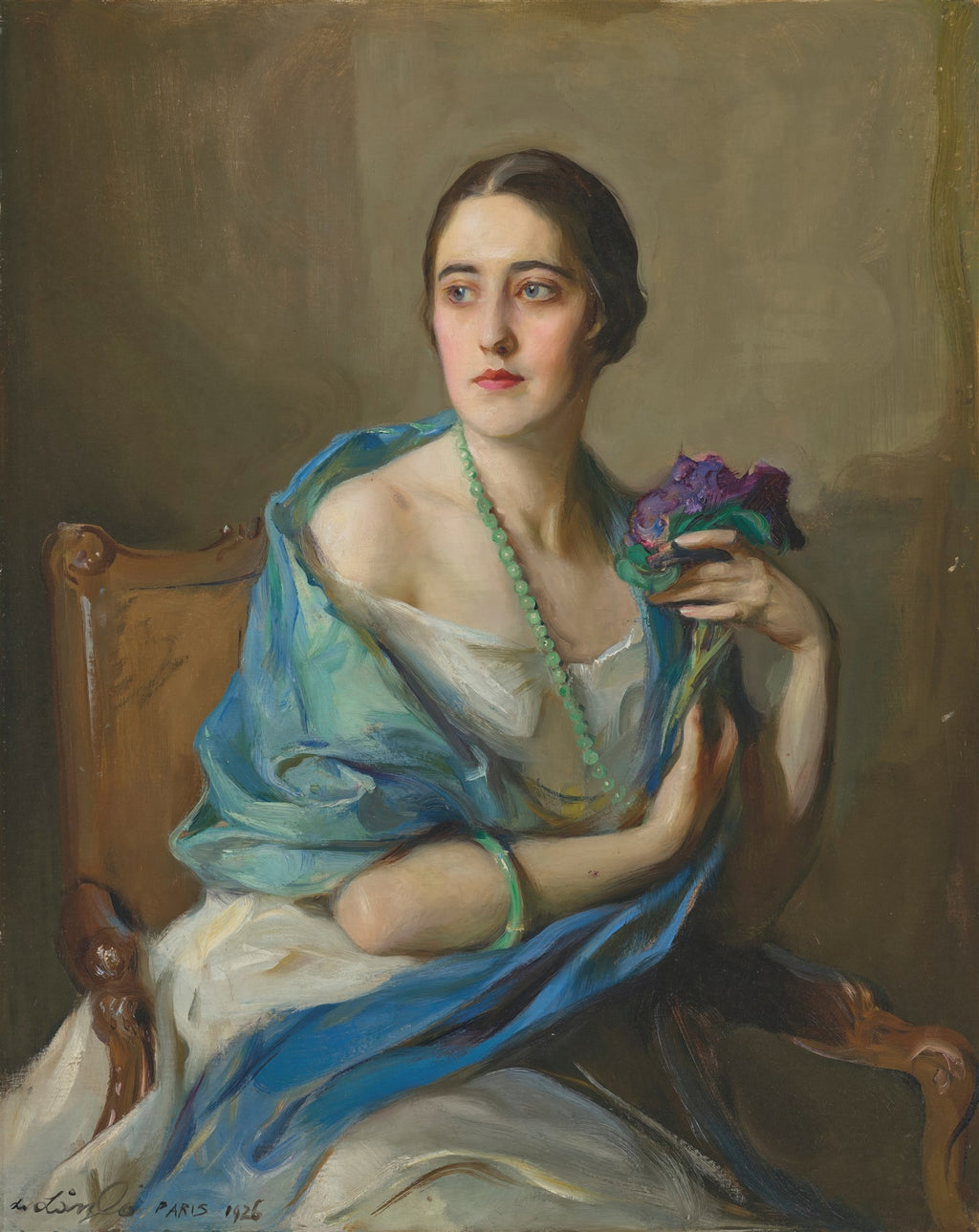 Retrato de uma mulher jovem e elegante, com cabelo escuro, usando um colar de contas verdes e um xale azul sobre um vestido branco, segurando flores violetas, sentada em uma cadeira de madeira, pintada por Philip Alexius de László.