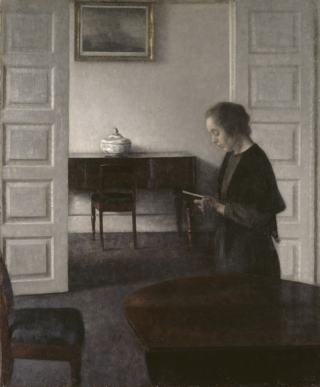 Quadro decorativo Interior com Mulher Lendo de Vilhelm Hammershøi. Uma mulher de perfil, vestida de preto, está lendo um livro em um ambiente minimalista e silencioso. As cores são predominantemente cinzas, marrons e brancas, com luz difusa entrando no cômodo.