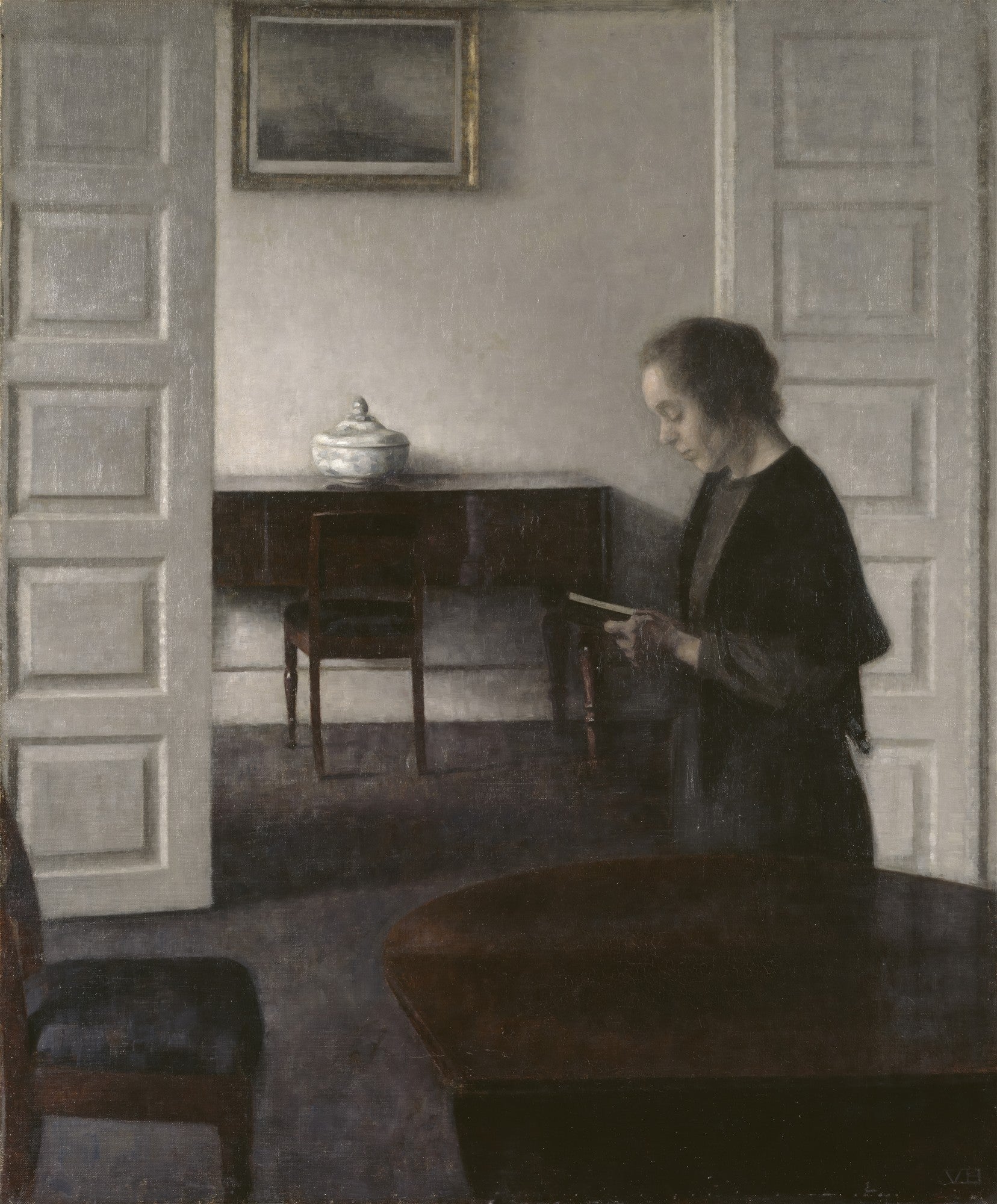 Quadro decorativo Interior com Mulher Lendo de Vilhelm Hammershøi. Uma mulher de perfil, vestida de preto, está lendo um livro em um ambiente minimalista e silencioso. As cores são predominantemente cinzas, marrons e brancas, com luz difusa entrando no cômodo.