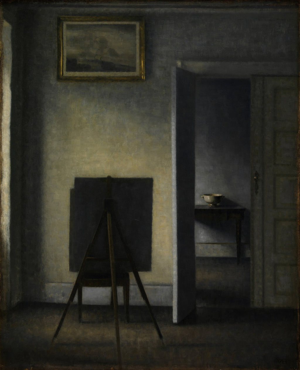 Quadro decorativo mostrando um interior minimalista e escuro com um cavalete de pintura em primeiro plano e uma porta aberta revelando outro cômodo à direita. A luz entra suavemente pela esquerda, iluminando a parede e o cavalete. Uma pequena moldura dourada com uma paisagem pendurada acima do cavalete. Obra de Vilhelm Hammershøi.