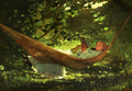 Quadro decorativo "A Rede" de Winslow Homer, retratando uma menina lendo em uma rede sob a sombra de árvores frondosas, com luz filtrada.